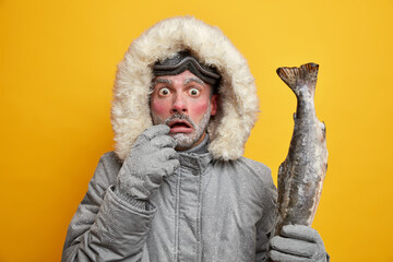 Outdoor winter activities and hobbies concept. Stupefied man with red frozen face stares bugged eyes holds caught big fish dressed in warm clothing has successful fishing isolated over yellow wall