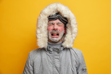 Dejected man cries and feels unhappy dressed in winter clothes feels very cold after snowboarding has red face covered with hoarfrost wears ski goggles isolated over yellow wall. Recreation concept - Powered by Adobe