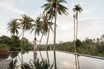 Summer vacation concept in tropics. Ypung woman in top and long skirt walking on the edge of infinity pool, surrounded with coconut palm trees and enjoy sunset time