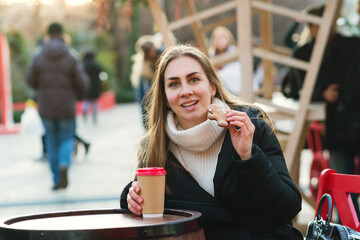 Young woman is walking at the Christmas market. Girl drinks takeaway coffee and eats gingerbread. Sweets. Christmas spirit. Merry Christmas and Happy Holidays