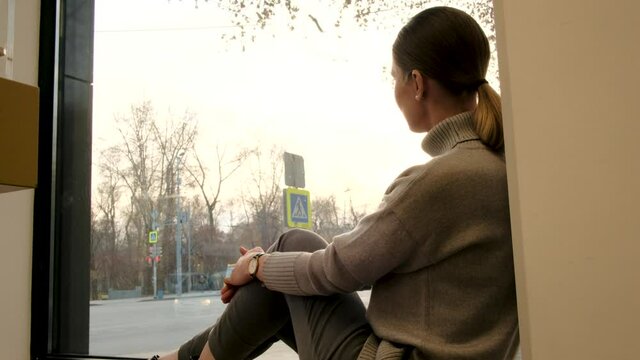 Brunette woman with short ponytail wearing cowl neck pullover looks at street sitting on windowsill in light room side view