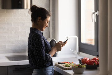 Happy young Indian woman read recipe on cellphone cooking healthy tasty dish at modern renovated home kitchen. Smiling ethnic female prepare delicious vegetarian salad use smartphone texting chatting.