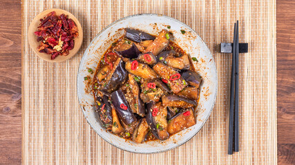 Korean spicy eggplant with spicy sauce and sesame seeds in a large bowl and chopsticks on a bamboo napkin on a rustic wooden table, top view