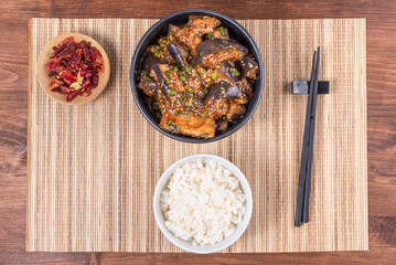Korean spicy eggplant with spicy sauce and sesame seeds in a bowl and a bowl of boiled rice on a bamboo napkin on a rustic wooden table, top view, close-up
