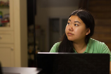 Asian teenage girl thinking and look aside in front of laptop computer while doing a homework or studying at home. Night scene, Education and online class concept.