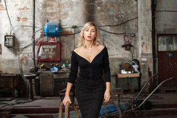 Fototapeta na wymiar Blonde woman wearing black dress holding a thick rope walking in an old spooky factory