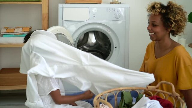 Young mother and daughter have fun and load washing machine in laundry at home spbd. American African woman and little girl talking with happy smiles and putting dirty clothes inside appliance. Modern