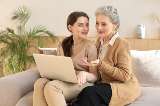 Happy elderly middle mother sitting on couch with her daughter, looking at laptop. Young woman showing video, photos to mommy, trusted relations. Family concept.