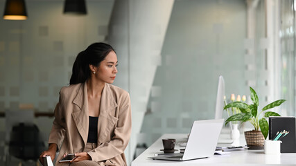 A young women entrepreneur in formal wear is sitting in front of laptop computer and looking out of window.