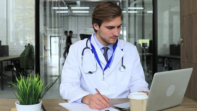 Doctor in white medical uniform with statoscope reading personal file of patient and writes data to laptop.