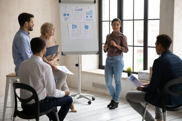 Young indian female speaker or coach present business project on flip chart to diverse employees in office. Ethnic businesswoman talk make whiteboard presentation for businesspeople at team training.