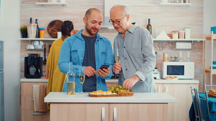 Young man showing his father how to use modern smartphone sitting in the kitchen and drinking a glass of white wine before family dinner. New lifestyle, using technology while women preparing the