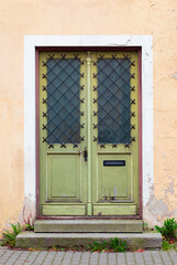 Fototapeta na wymiar Green retro double doors with glass panels, diagonal iron bars, a letterbox, small stairs and cracks in paint