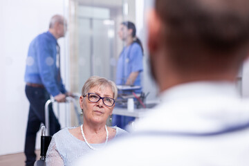 Fototapeta premium Handicapped woman in wheelchair at doctor appointment. Recovery treatment, help for disabled people. Elderly assistance. Old man with walking frame on hospital corridor.