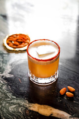 An orange cocktail in a rocks glass with a big ice cube, paprica crust rim, served with almonds