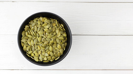 Pumpkin seeds in black bowl on white wooden table. Top view. Copyspace.
