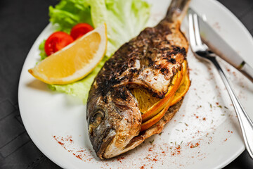 A close-up of grilled dorado fish stuffed with orange, garnished with lettuce, lemon and cherry tomato, knife and fork