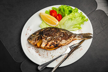 Grilled dorado fish stuffed with orange, garnished with lettuce, lemon and cherry tomato, knife and fork, top view