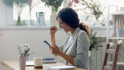 Excited young Caucasian woman sit at desk at home office look at smartphone screen feel euphoric with good news on gadget. Overjoyed millennial female triumph winning online lottery on cellphone.