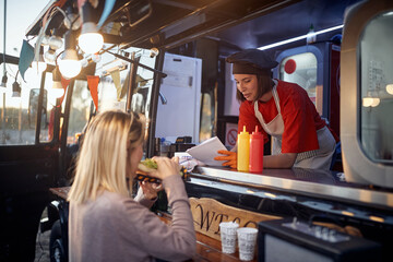 young female customer eating sandwich in front of fast food service, giving suggestions to a beautiful employee