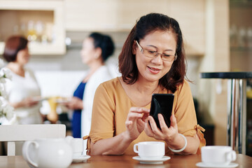 Portrait of mature Asian woman in glases drinking tea and answering messages on smartphone