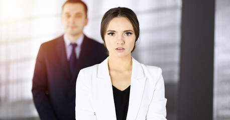 Young intelligent businesswoman is standing straight in a sunny modern office with her colleague at the background. Concept of successful business