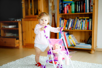 Cute adorable baby girl making first steps with doll carriage. Beautiful toddler child pushing stroller with toy at home. Happy daughter learning walking and standing