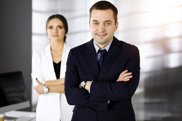 Portrait of a friendly middle aged businessman in a dark blue suit, standing with crossed arms together with a colleague in a sunny modern office. Modern business concept