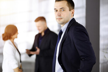 Portrait of a self-confident middle aged businessman in a blue suit, standing in a sunny modern office with his colleagues at the background. Concept of business success