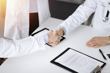 Unknown doctors are shaking their hands finishing discussion about patient's diagnosis, sitting at the desk and using a clipboard, close-up. Medical help, insurance in health care