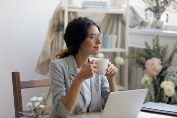 Smiling young Caucasian businesswoman in glasses drink coffee enjoy morning in home office. Happy female fashion designer stylist look in distance dreaming thinking, work in cozy creative workplace.
