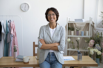 Portrait of successful happy young Caucasian female tailor or stylist posing at desk in modern home office or atelier. Smiling confident woman fashion designer feel motivated inspired in boutique.