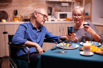 Disabled man using smartphone in kitchen while dining with mature wife. Scrolling showing photos. Imobilized handicapped senior husband scolling on phone enjoying the festive meal.