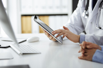 Unknown male doctor and patient woman discussing current health examination while sitting in clinic and using tablet computer, closeup of hands. Medicine and healthcare concept