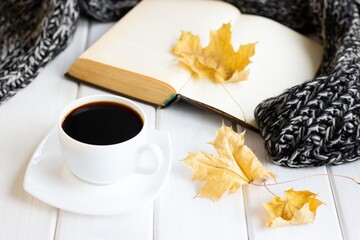 Yellow autumn leaves, coffee cup, warm knitted scarf with opened old book on white wooden boards. Cozy home reading, hygge concept