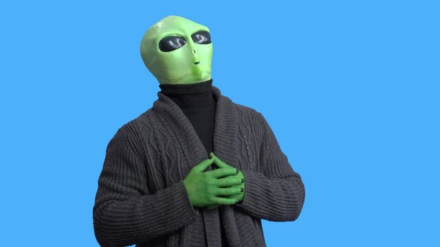 Man in alien costume, warm knitted jacket, with funny themed mug in his hand laughs, writhes with laughter, against a blue chroma key. Preparation for Halloween party.
