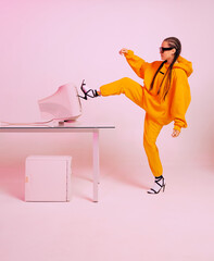 Stylish female office worker in sunglasses and hoodie kicks obsolete computer monitor. Colored Neon light
