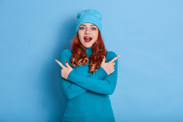 Good looking female isolated over blue background, pointing with both hands to both sides, keeping mouth open wide, being surprised and astonished about excited news, wearing jumper and cap.