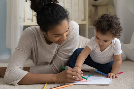 Caring happy African American mom lying on floor at home drawing with little baby infant. Loving smiling biracial mother feel playful paint engaged in funny activity with small toddler child.