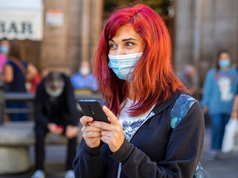 Mujer joven con pelo rojo usando su teléfono móvil vistiendo mascarilla para respetar las medidas de seguridad y distanciamiento social de la pandemia del coronavirus