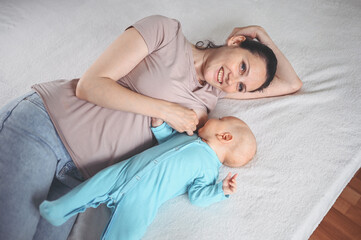 Young mother lies with newborn infant baby in blue jumpsuit on bed and breastfeeds him breast milk