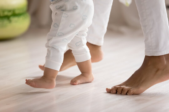 Crop close up of loving mother hold little biracial child hands learn walking at home on wooden floor. Small cute ethnic baby toddler make first steps with mom support and care. Childcare concept.