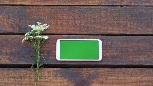 Closeup top view 4k video of white smartphone with empty blank green screen. Cell laying on brown vintage wooden tabel background. Woman scrolling invisible virtual pages.