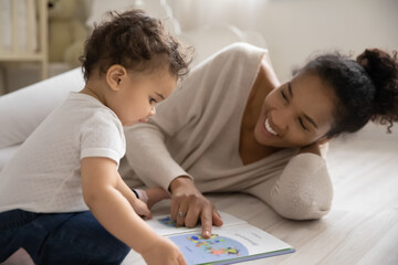 Smiling loving African American mom lying on warm floor in children room reading book with curious biracial baby infant. Happy caring ethnic mother engaged in funny learning activity with small child. - Powered by Adobe