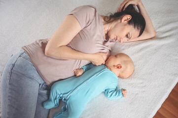 Young mother lies with newborn infant baby in blue jumpsuit on bed and breastfeeds him breast milk