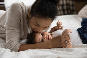 Portrait of loving happy young african American mother lying on bed with little baby infant kiss...