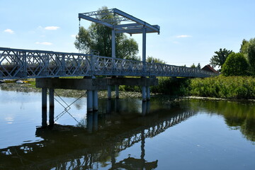 Close up on a metal pedestrian and car bridge connecting two river banks or coasts with one of the coasts being overgrown with shrubs, reeds, and other types of flora seen on a sunny summer day