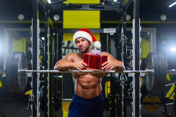 Young muscular attractive Santa Claus in Christmas hat is holding a gift leaning on a barbell in a gym, front view