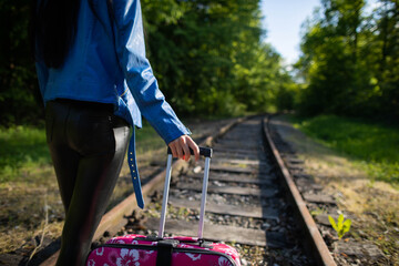 A close-up view of a teenage girl holding a pink travel bag by a retractable plastic handle while walking along the railroad tracks.