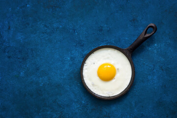 Fried egg in a small cast iron skillet. Blue grunge background. Modern minimalist food. Top view, flat lay.